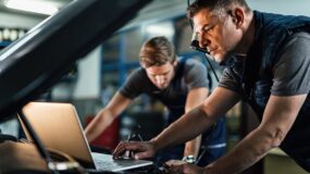 Two men in a garage working on a vehicle, one man stands in the background looking down at something and the other stands looking at a laptop