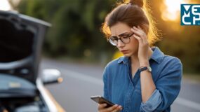 A woman with glasses looking down at a cellphone in her hand while in the background, there is a vehicle with its hood open